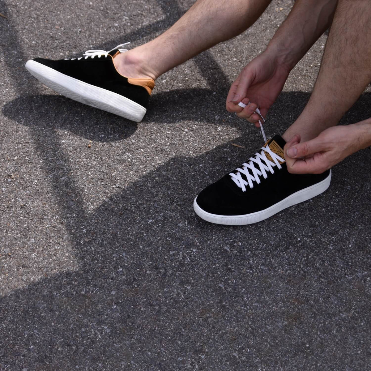 Man with black Hardwick sneakers on tying up his left shoe.