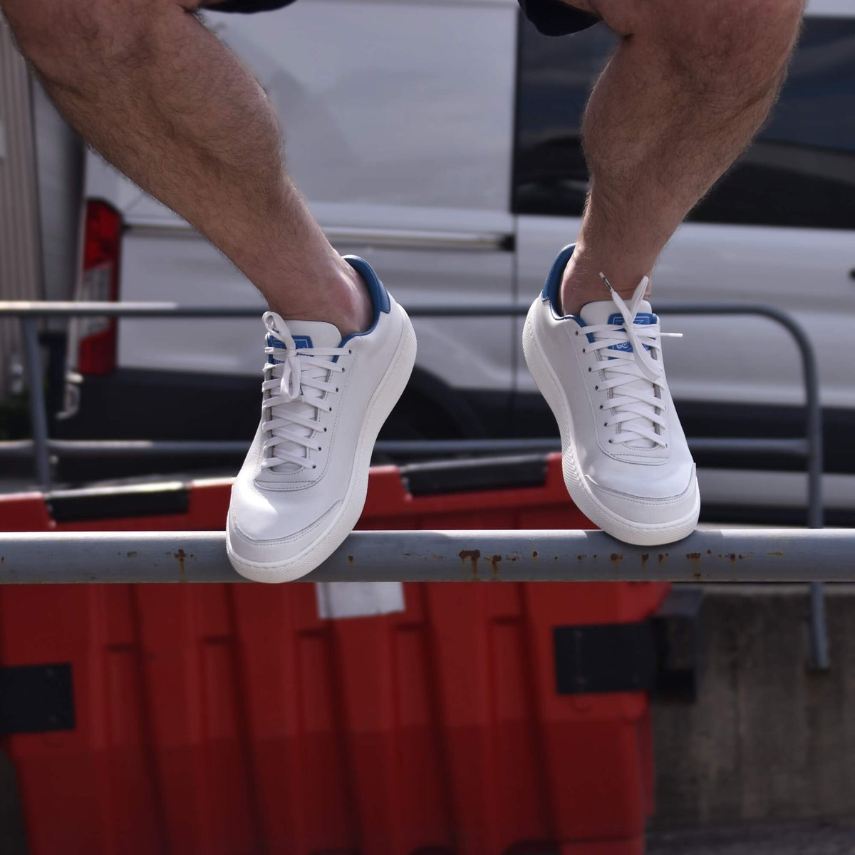 Man sat on a rail wearing white Hardwick sneakers seen from the shins down